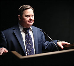 Mark Hublar poses in front of a podium as he would at any of his public speaking events to advocate for employment of those with disabilities and to share the message that “we’re all different, but the same.” (Submitted photo)